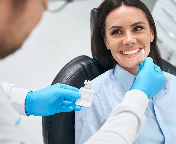 Woman in blue shirt in dental chair smiling at dentist holding mini shade guide